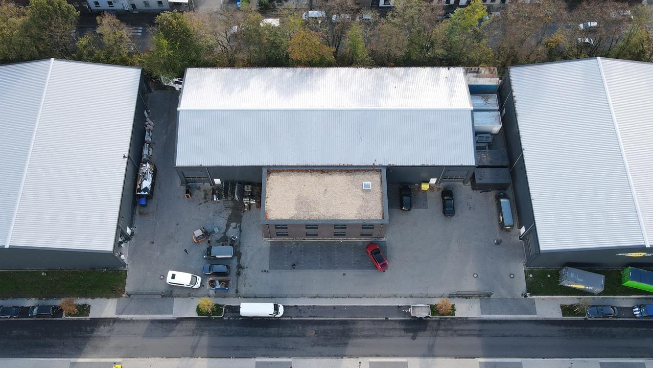 Luftaufnahme eines Gewerbegebäudes mit Flachdach und angrenzendem Parkplatz, umgeben von weiteren Hallen und einer Straße im Vordergrund